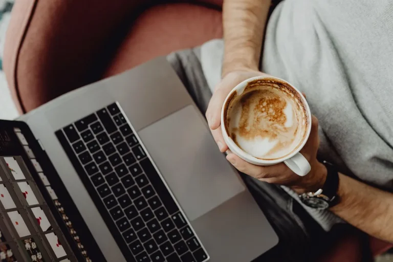 Een close-up van iemand die een kop cappuccino vasthoudt met een laptop op schoot, wat een gezellig moment van ontspanning suggereert.